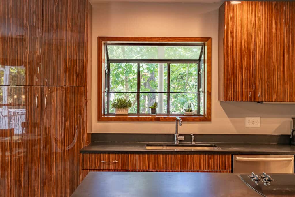 A close-up view of a modern kitchen sink and window framed with rich wooden details, overlooking a lush green outdoor area.