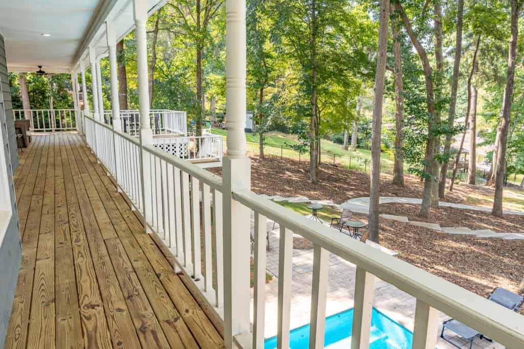 A spacious wooden wraparound porch with white railings, overlooking a landscaped backyard with trees and a pool.