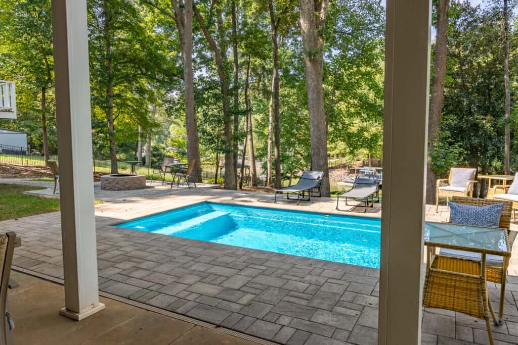 View of a serene backyard featuring a sparkling pool, fire pit, and lush wooded surroundings seen from the covered patio area.
